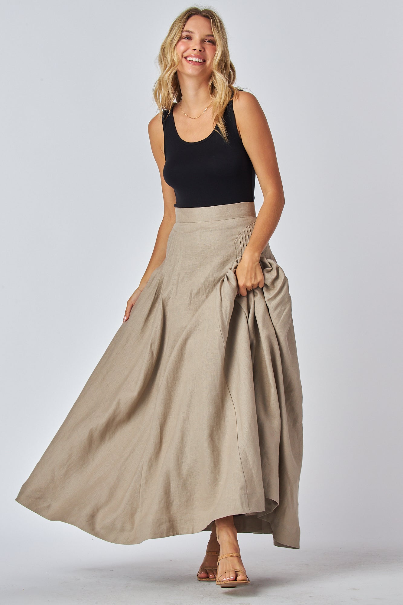 A model wearing a flowing maxi skirt in a light tan color with a high waistband and side pleated design, paired with a black tank top.