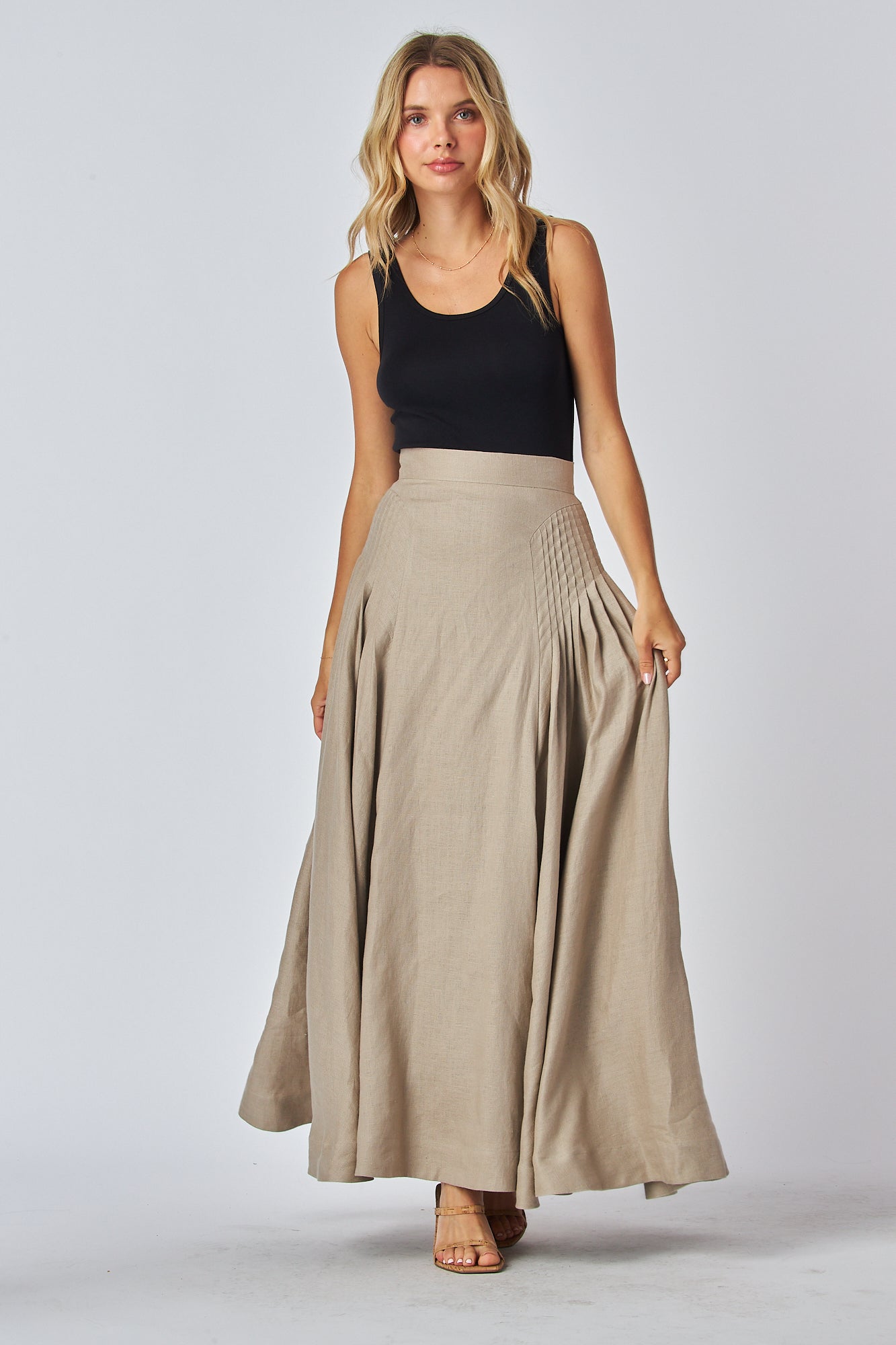 A model wearing a flowing maxi skirt in a light tan color with a high waistband and side pleated design, paired with a black tank top.