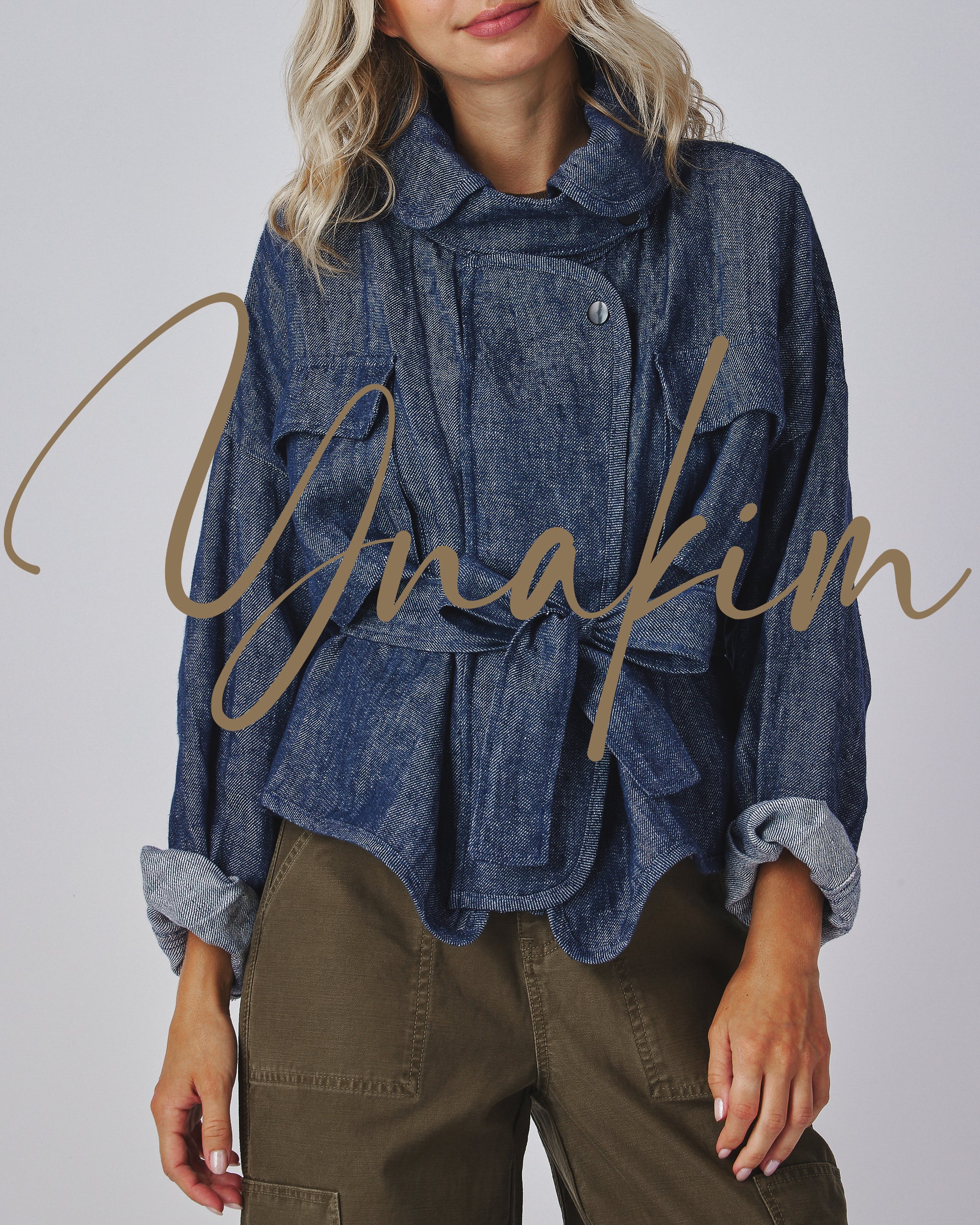 A blonde woman models a blue herringbone jacket with a wide lapel collar, a front tie-belt, and two patch pockets on the chest.